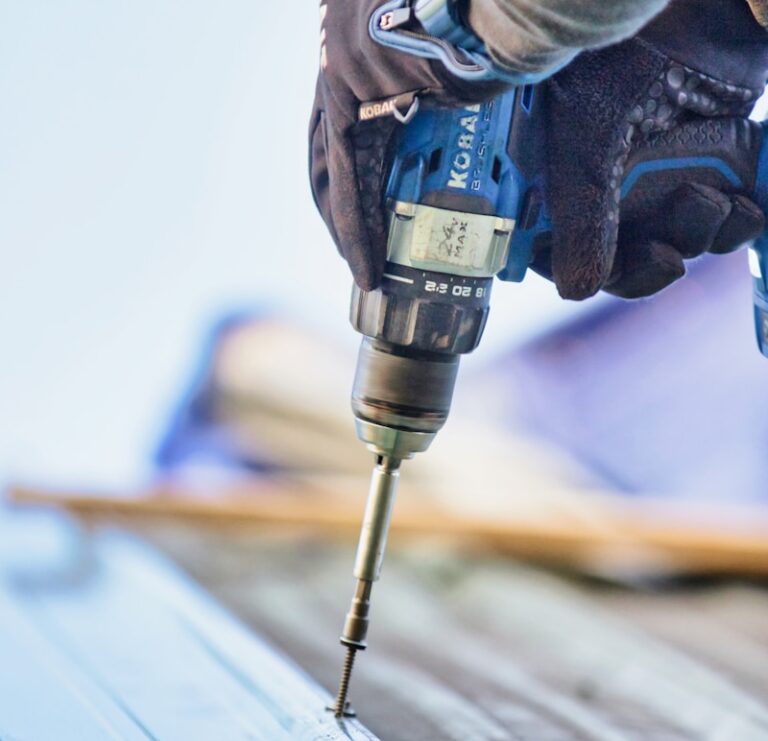 A roofing contractor in Lyndhurst, wearing gloves, uses a cordless power drill to drive a screw into a surface. The focus is on the hands, drill, and screw against a blurred background.
