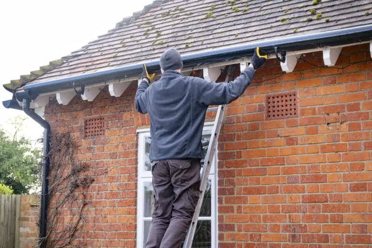 A person stands on a ladder, cleaning or inspecting the gutters of a brick house—a typical scene for gutter service in Lyndhurst. They wear a beanie, gloves, and dark jacket. The sloped roof has some moss and there's a white-framed window below.