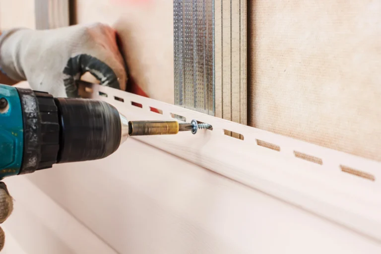 A Siding Contractor in Lyndhurst, wearing gloves, uses a power drill to attach a screw through a vinyl siding panel onto a wall with metal studs.