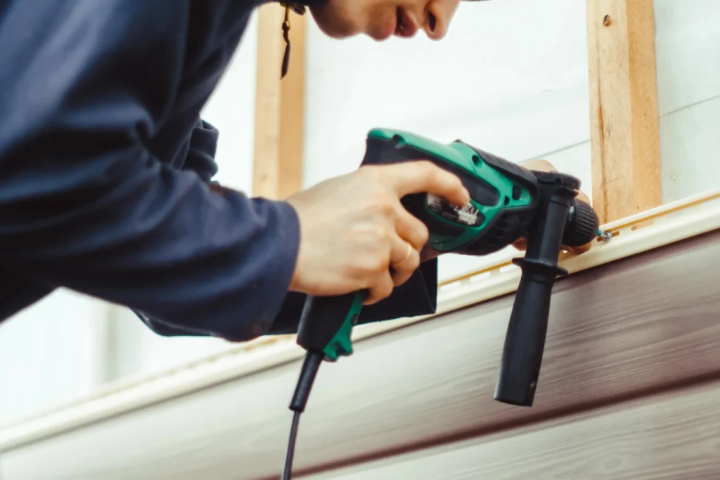 A focused Siding Contractor in Lyndhurst uses a green and black electric drill to secure siding to a wall, carefully positioning the tool near the wooden framing.