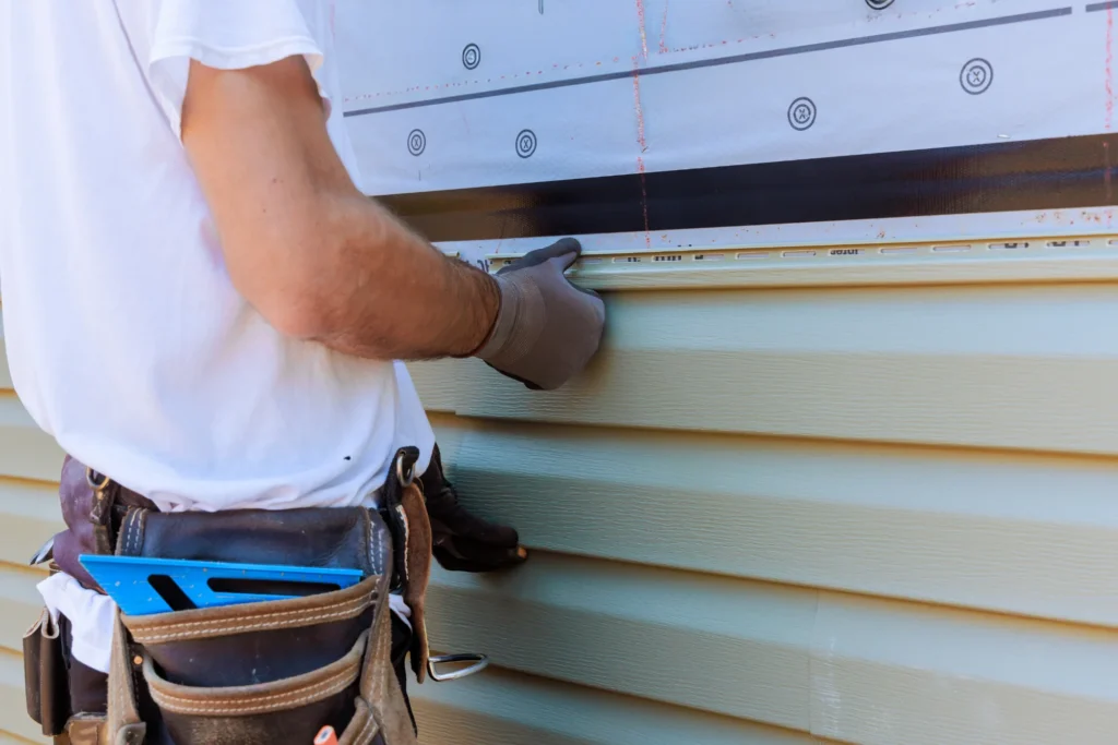 A construction worker, likely a Siding Contractor in Lyndhurst, wears gloves and a tool belt while installing light green vinyl siding on a house exterior, carefully using a measuring tool to ensure proper alignment.
