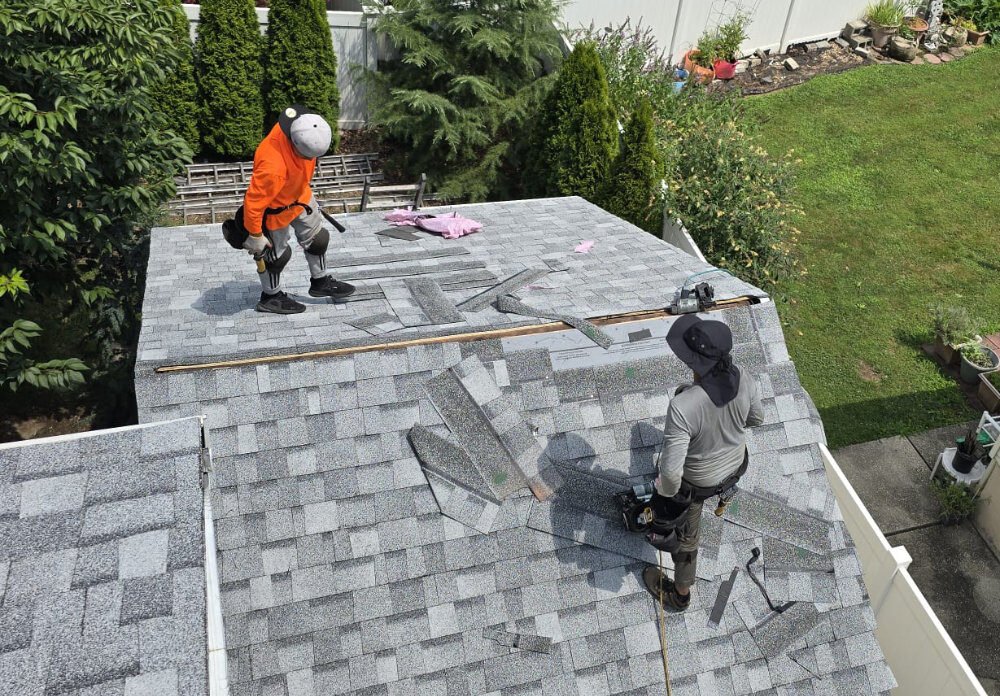 Two workers from United Pro Restoration in safety gear are installing gray shingles on a sloped roof. Roofing materials and tools are scattered around them. Trees, grass, and a white fence are visible in the background.