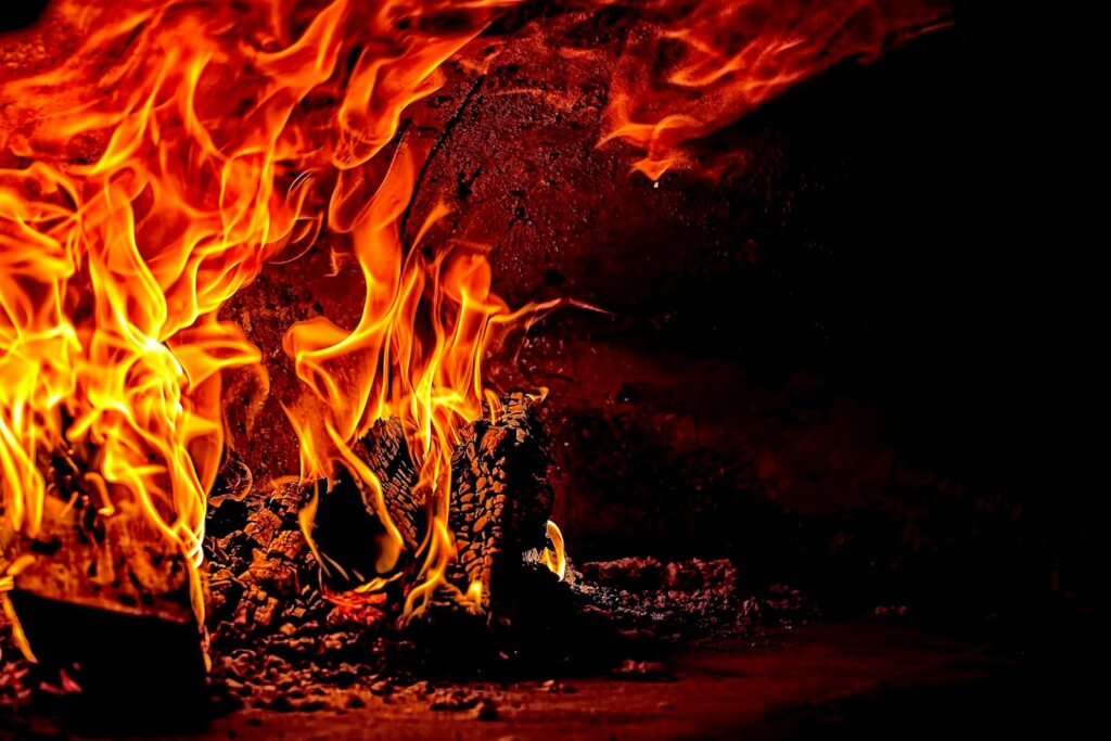 Bright orange and yellow flames burn intensely over wood and coals inside a dark, rustic fireplace, casting dramatic light and shadows on the rough stone behind—reminding some of the need for expert Fire Damage Restoration Service in Lyndhurst.