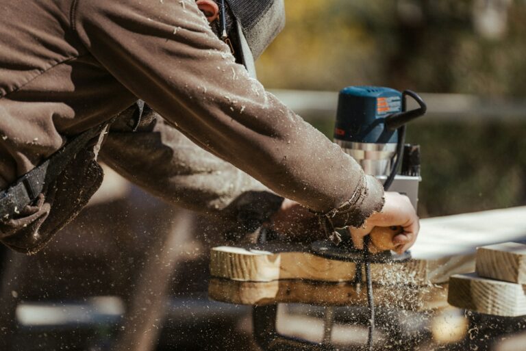 A General Contractor in Lyndhurst uses a handheld power tool to shape or sand a wooden board, with wood shavings flying in the air, suggesting active woodworking outdoors.